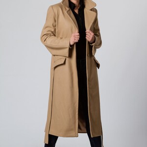 May include: A long camel-colored coat with a black button-up shirt underneath. The coat has a large collar and two pockets. The person is wearing brown boots.
