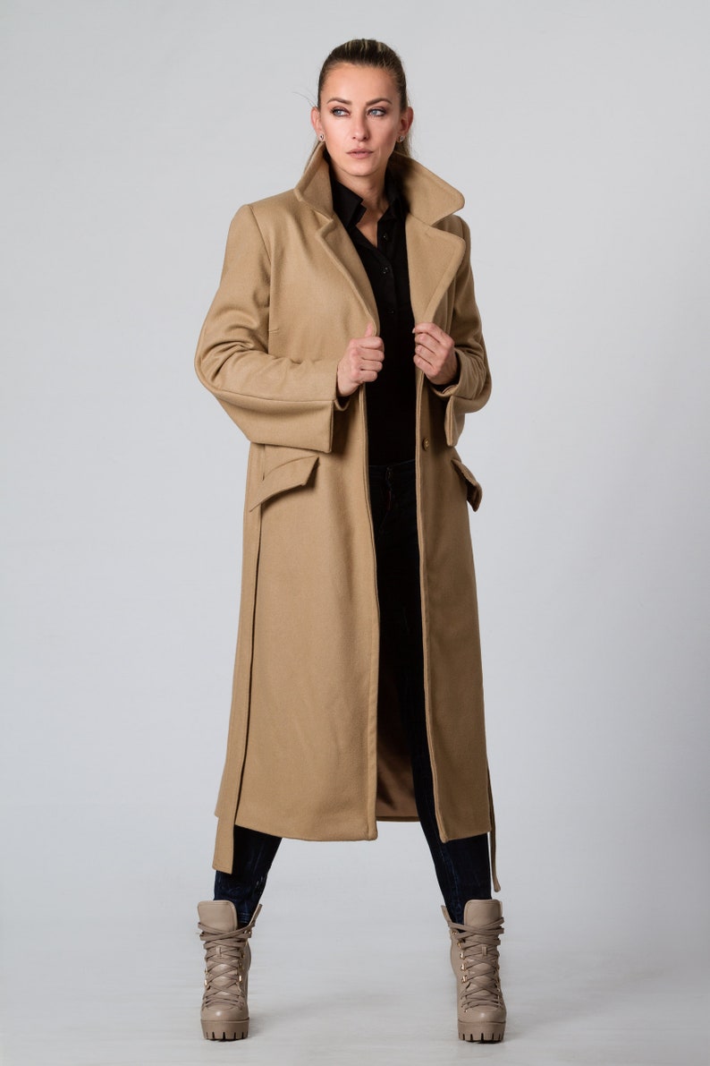 May include: A long camel-colored coat with a black button-up shirt underneath. The coat has a large collar and two pockets. The person is wearing brown boots.