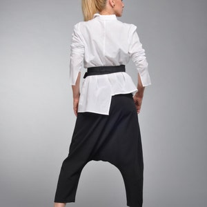 May include: A woman wearing a white button-down shirt with a black belt and black pants. The pants have a unique design with a gathered crotch.