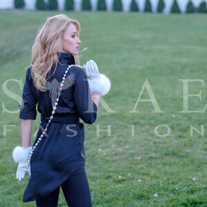 May include: A woman wearing a black shirt with a bird design and a pearl necklace. She is wearing white fur gloves and is standing in a grassy field.