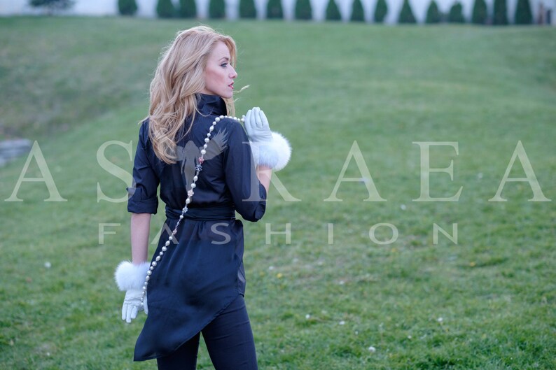 May include: A woman wearing a black shirt with a bird design and a pearl necklace. She is wearing white fur gloves and is standing in a grassy field.