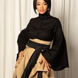 Op de afbeelding: Een zwarte top met hoge kraag en knoopsluiting, gecombineerd met een beige rok en een brede zwarte riem. De outfit heeft een modern, elegant ontwerp, geschikt voor formele gelegenheden.
