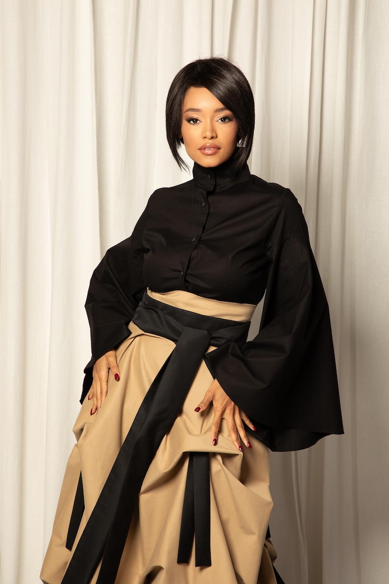 Op de afbeelding: Een zwarte top met hoge kraag en knoopsluiting, gecombineerd met een beige rok en een brede zwarte riem. De outfit heeft een modern, elegant ontwerp, geschikt voor formele gelegenheden.