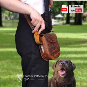 Peut inclure: Un sac à bandoulière en cuir marron avec une fermeture à rabat et une boucle en métal. Le sac est porté par une personne vêtue d'une tenue noire. Un chien brun est assis dans l'herbe au premier plan.