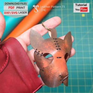 Könnte beinhalten: Ein handgefertigter Leder-Katzenmasken-Schlüsselanhänger mit einem Used-Look-Finish in Braun und Orange. Die Maske hat genähte Details, Augenlöcher und einen Schlüsselring. Das Bild enthält auch den Text "Download Files PDF Print DXF/SVG Laser" und "Tutorial You Tube".