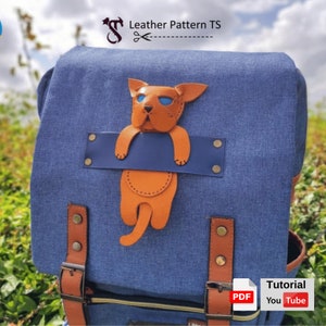 Könnte beinhalten: Ein blauer Canvas-Rucksack mit einem braunen Leder-Katzen-Applik. Die Katze hängt an einem blauen Lederband und hat orangefarbenes Fell und blaue Augen. Der Rucksack hat braune Lederriemen und Schnallen. Der Text "PDF Tutorial YouTube" ist in der unteren rechten Ecke des Bildes sichtbar.