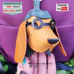 Può includere: Un portachiavi a forma di cane in pelle fatto a mano, con occhiali rotondi neri e un cappello verde. Il cane è marrone chiaro con orecchie marroni e un naso nero. L'immagine include il testo "DOWNLOAD FILES PDF PRINT DXF/SVG LASER" e "Tutorial You Tube".