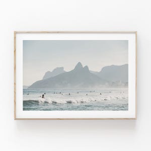 Puede incluir: Una impresión enmarcada que presenta una escena costera con surfistas en el océano. La imagen muestra una cordillera en el fondo, con una paleta de colores azul claro y gris. La impresión está encerrada en un marco de madera de color claro.