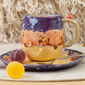May include: A handmade ceramic mug with a matching saucer. The mug is a mix of purple, orange, and yellow with a unique handle and a small red accent. The set includes a spoon and colorful candies.