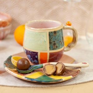 Pode incluir: Uma caneca de cerâmica colorida com um design de patchwork, pousada em um pires correspondente com uma colher pequena. A caneca apresenta quadrados verdes, azuis, laranja e roxos. Uma trufa de chocolate está na colher.