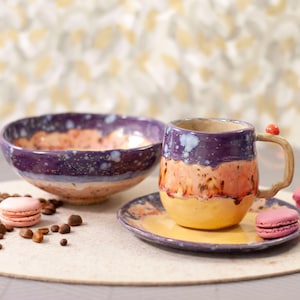 Puede incluir: Un juego de cuenco, taza y platillo de cerámica hechos a mano. El cuenco y la taza son morados, naranjas y amarillos con un esmalte moteado. La taza tiene un asa única y un pequeño acento rojo. Macarrones rosas y granos de café están dispuestos.