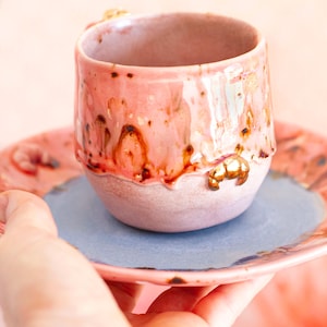 May include: A handmade ceramic cup and saucer set. The cup is pink with brown and gold accents, and a gold-colored handle. The saucer is pink with a blue center. The set is held in a hand.