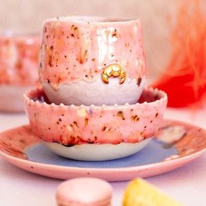 Op de afbeelding: Een roze keramische kop en schotel set met een gouden krab accent. De kop en kom hebben een gespikkeld ontwerp en een witte basis. De set bevat een kleine kom en een bord met een blauw centrum.