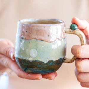 Op de afbeelding: Een handgemaakte keramische mok met een uniek, gelaagd ontwerp. De mok heeft banden in bruin, groen en blauw, met een textuur en organische uitstraling. De handgreep heeft een decoratieve groene kraal.