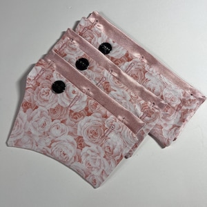 May include: Four small, rectangular pouches with a floral rose pattern in shades of pink and white. Each pouch has a pink satin border and a black button closure. The pouches are stacked slightly, revealing the rose pattern.