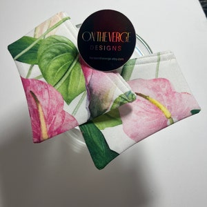 May include: Two white fabric pouches with a pink and green floral print. The pouches are folded and stacked on top of each other. A black and gold sticker with the text "ON THE VERGE DESIGNS" and the website "horseontheverge.etsy.com" is on top of the pouches.