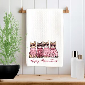 Peut inclure: Serviette de cuisine blanche avec quatre chats portant des pulls roses et des lunettes de soleil en forme de c&oelig;ur. Le texte "Happy Meownentine's" est imprim&eacute; sous les chats.