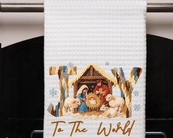 May include: White waffle-weave kitchen towel with the word "JOY" and the phrase "To The World" embroidered in brown. The center features a Nativity scene with Mary, Joseph, baby Jesus, and sheep. Snowflakes are scattered around the letters.