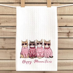 Peut inclure: Une serviette de cuisine blanche avec une illustration rose et brune de quatre chats portant des lunettes en forme de c&oelig;ur et des pulls roses. Le texte "Happy Meowentines" est imprim&eacute; sous les chats.