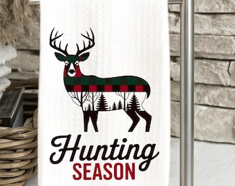 May include: White kitchen towel with a deer silhouette, featuring a red and green plaid pattern. The deer's body incorporates a forest design. The words "Hunting SEASON" are written in a script font.