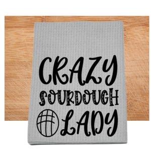 Puede incluir: Una toalla de cocina gris con el texto "CRAZY SOURDOUGH LADY" en letras negras. Una ilustración en blanco y negro de una hogaza de pan está a la izquierda del texto.