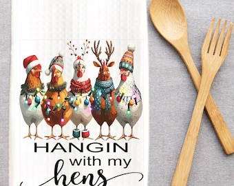 Peut inclure: Serviette de cuisine blanche avec un motif festif de cinq poulets ornés de chapeaux de fête, d'écharpes et de guirlandes lumineuses. Le texte "HANGIN with my hens" est imprimé en dessous. Cuillère et fourchette en bois à droite.