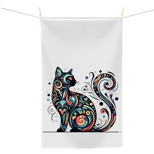 May include: A white, textured towel with a colorful cat design. The cat is black with intricate patterns in shades of blue, orange, yellow, and green. The towel is hanging from a clothesline.