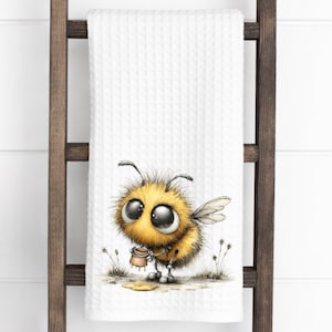May include: A white waffle-weave kitchen towel featuring a central illustration of a fuzzy, round yellow bee with large black eyes. The bee is holding a small brown honey pot, with a small puddle of spilled honey on the ground near its feet.