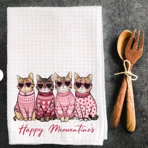 Peut inclure: Serviette de cuisine blanche avec un motif rose et blanc de quatre chats portant des lunettes en forme de c&oelig;ur et des pulls roses. Le texte "Happy Meowentines" est imprim&eacute; sous les chats.