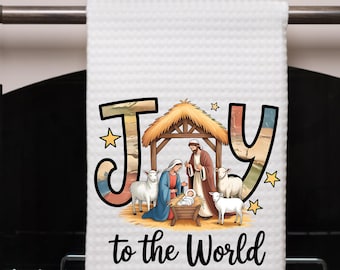 May include: White waffle-weave kitchen towel with a colorful illustration of the Nativity scene. The word "Joy" is written in large letters, and the phrase "to the World" is below the illustration. The towel is hanging on a metal bar.