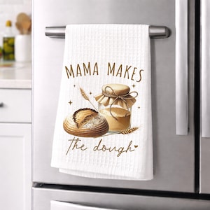 Puede incluir: Toalla de cocina blanca con tejido de gofres con una ilustración de acuarela de pan, un tarro de harina y espigas de trigo. El texto "MAMA MAKES the dough" está impreso en escritura marrón. La toalla cuelga de un tirador de nevera de acero inoxidable.