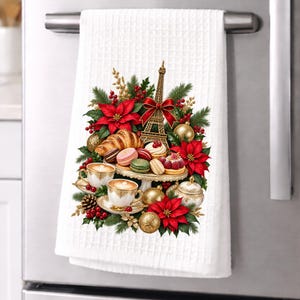 May include: A white waffle-weave kitchen towel featuring a festive holiday illustration of the Eiffel Tower, red poinsettias, pine branches, gold ornaments, a croissant, colorful macarons, and two cups of coffee on a tiered serving tray.