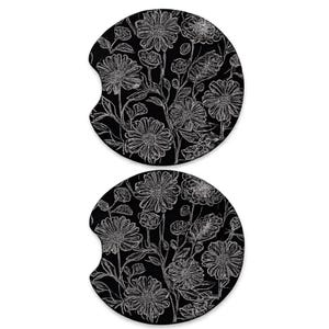 May include: Two black car coasters with a white floral design. The coasters have a semi-circular shape with a notch for easy removal. The floral pattern includes daisies and other flowers with stems and leaves.