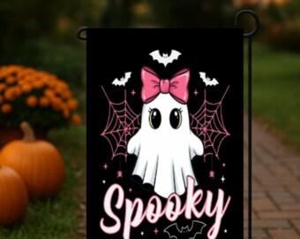 May include: A black Halloween garden flag featuring a cartoon ghost with a pink bow, spiderwebs, and the word "Spooky" in pink script. Bats are also included in the design. The flag is displayed outdoors with pumpkins and flowers in the background.