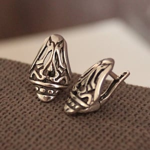 May include: A pair of silver-toned earrings with an ornate, openwork design. The earrings have a teardrop shape with intricate patterns and a hinged closure. The jewelry is displayed on a textured, brown surface.