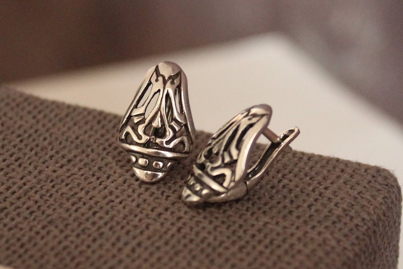 May include: A pair of silver-toned earrings with an ornate, openwork design. The earrings have a teardrop shape with intricate patterns and a hinged closure. The jewelry is displayed on a textured, brown surface.