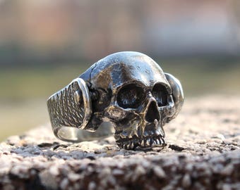 Può includere: Un anello in argento a forma di teschio con una fascia testurizzata e due accenti decorativi sui lati. Il teschio è dettagliato con orbite, un naso e una bocca.