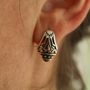 May include: Close-up of a silver earring with an ornate, openwork design. The earring has a teardrop shape with a detailed pattern and a small, rounded base. The background is blurred, focusing on the jewelry.