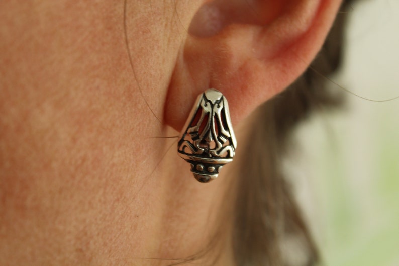 May include: Close-up of a silver earring with an ornate, openwork design. The earring has a teardrop shape with a detailed pattern and a small, rounded base. The background is blurred, focusing on the jewelry.