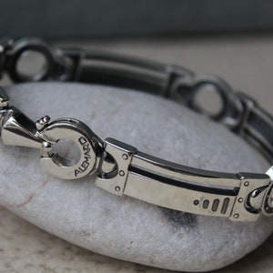 May include: A silver-toned bracelet with a unique, industrial design. The bracelet features a series of interconnected links with circular and rectangular elements. The clasp is engraved with the word "ALEMARD".
