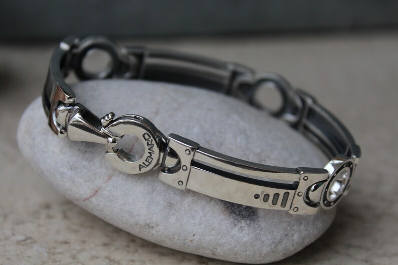 May include: A silver-toned bracelet with a unique, industrial design. The bracelet features a series of interconnected links with circular and rectangular elements. The clasp is engraved with the word "ALEMARD".