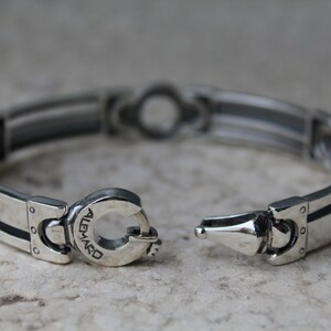 May include: Silver bracelet with a unique clasp design. The bracelet features a series of rectangular links with black accents and a central decorative element. The clasp is engraved with the word "ALEMARD".