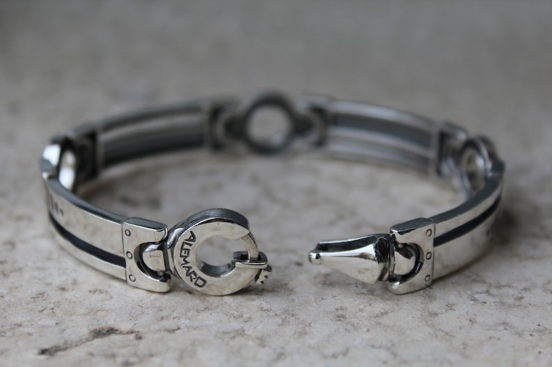May include: Silver bracelet with a unique clasp design. The bracelet features a series of rectangular links with black accents and a central decorative element. The clasp is engraved with the word "ALEMARD".