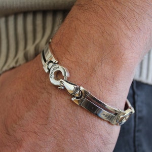 May include: A silver bracelet with a unique clasp design is shown on a person's wrist. The bracelet has a polished finish with a geometric pattern. The clasp is circular and has the word "ALVARO" engraved on it.