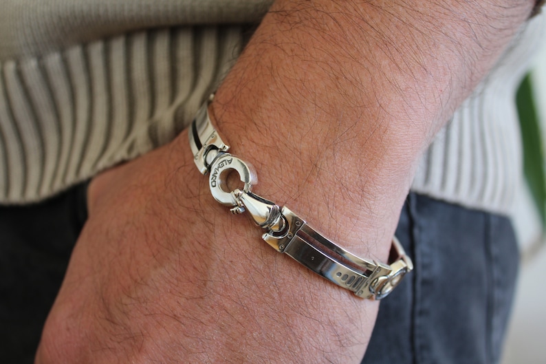 May include: A silver bracelet with a unique clasp design is shown on a person's wrist. The bracelet has a polished finish with a geometric pattern. The clasp is circular and has the word "ALVARO" engraved on it.