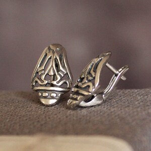 May include: A pair of silver-colored earrings with an openwork design. Each earring has a teardrop shape with intricate cut-out patterns and a hinged clasp. The earrings are displayed on a textured, gray surface.