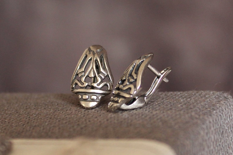 May include: A pair of silver-colored earrings with an openwork design. Each earring has a teardrop shape with intricate cut-out patterns and a hinged clasp. The earrings are displayed on a textured, gray surface.