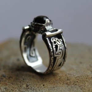 May include: A silver ring with a dark, possibly black, gemstone. The ring has an ornate design with etched patterns and a textured band. The gemstone is set in a detailed bezel. The ring is displayed on a neutral surface.
