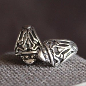May include: Two silver rings with intricate openwork designs. The rings have a detailed pattern of swirling lines and shapes, with a slightly domed top. The rings are displayed on a textured gray surface.