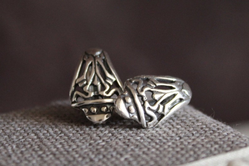 May include: Two silver rings with intricate openwork designs. The rings have a detailed pattern of swirling lines and shapes, with a slightly domed top. The rings are displayed on a textured gray surface.